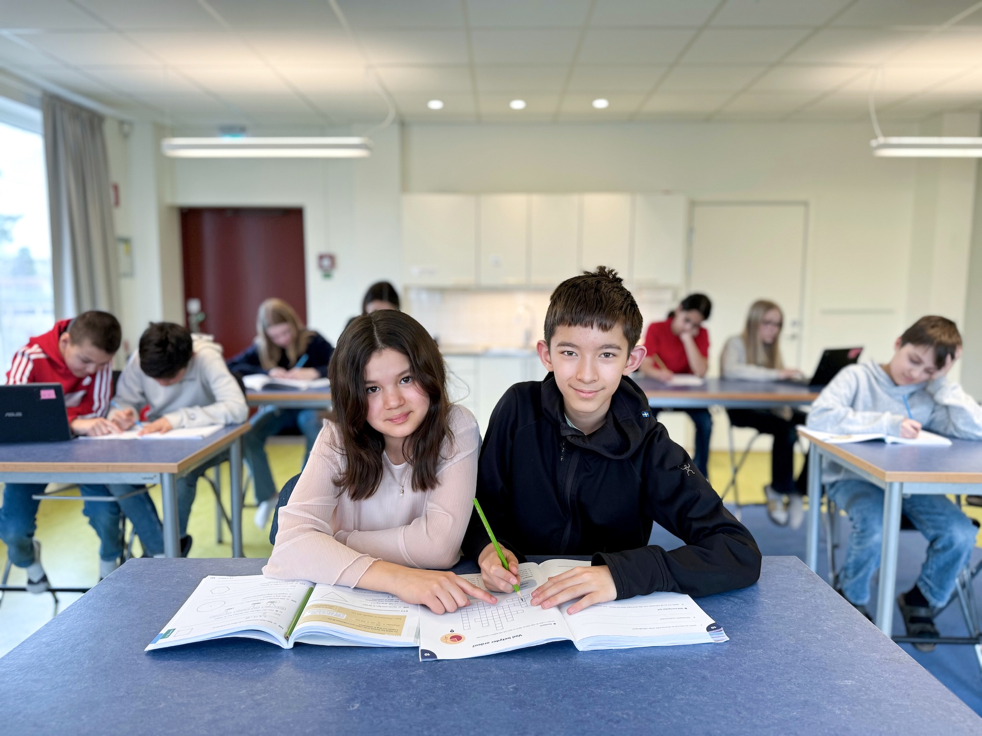 två elever i klassrum med böcker framför sig, som tittar in i kameran